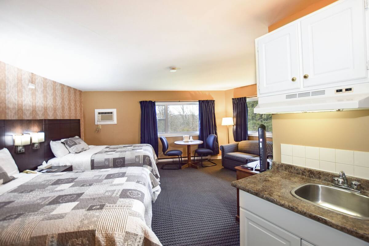 Habitación de hotel con dos camas, una pequeña mesa de comedor, un sofá, un área de cocina con un lavamanos y una ventana con cortinas.