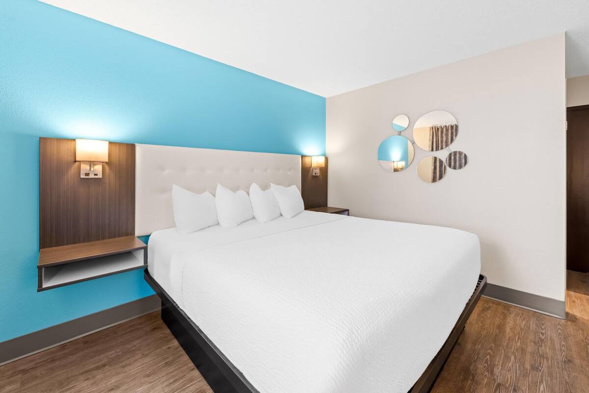 A modern hotel room with a large bed, a wooden nightstand, a wall-mounted lamp, and a decorative mirror.