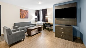 A modern hotel room with a grey sofa, a wooden coffee table, a desk with a chair, a flat-screen TV, and a wardrobe.