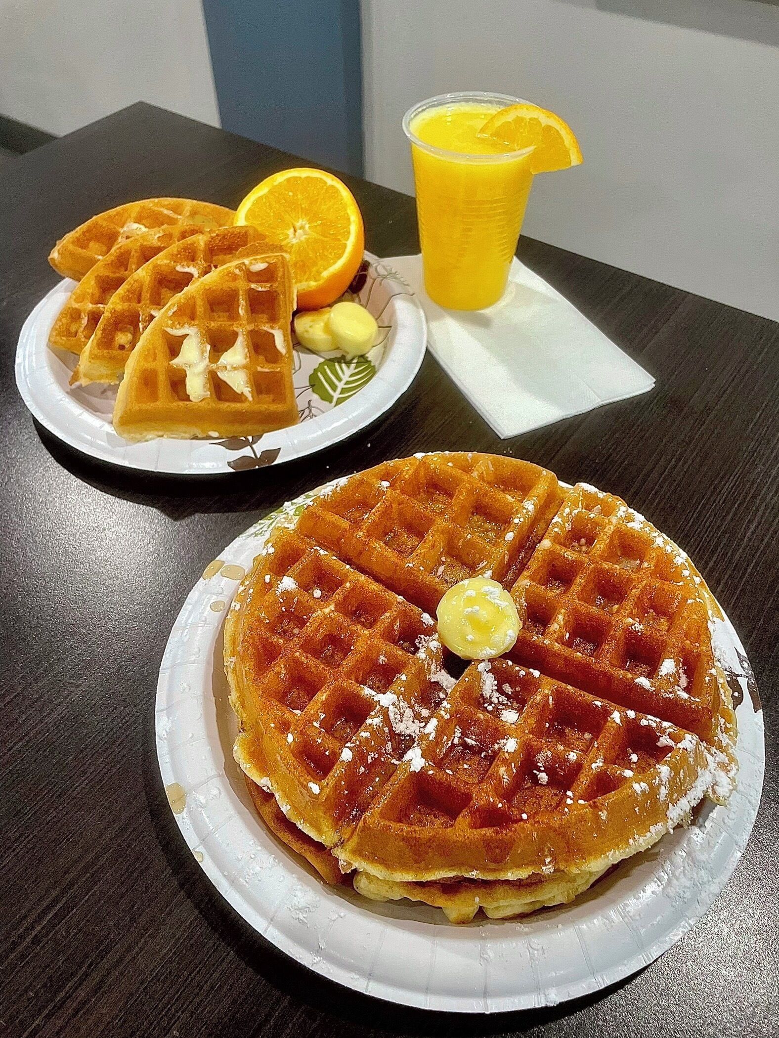 A plate of waffles with powdered sugar and a slice of lemon on a white plate, another plate with waffles and orange slices, and a glass of orange juice on a table.