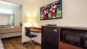 A hotel room with a television mounted on the wall displaying a sports game, a desk with a lamp, a chair, and a refrigerator.