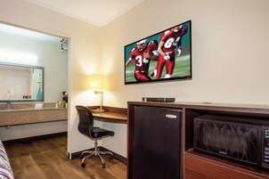 Habitación de hotel con una televisión en la pared mostrando un partido deportivo, un escritorio con una lámpara, una silla y un refrigerador.