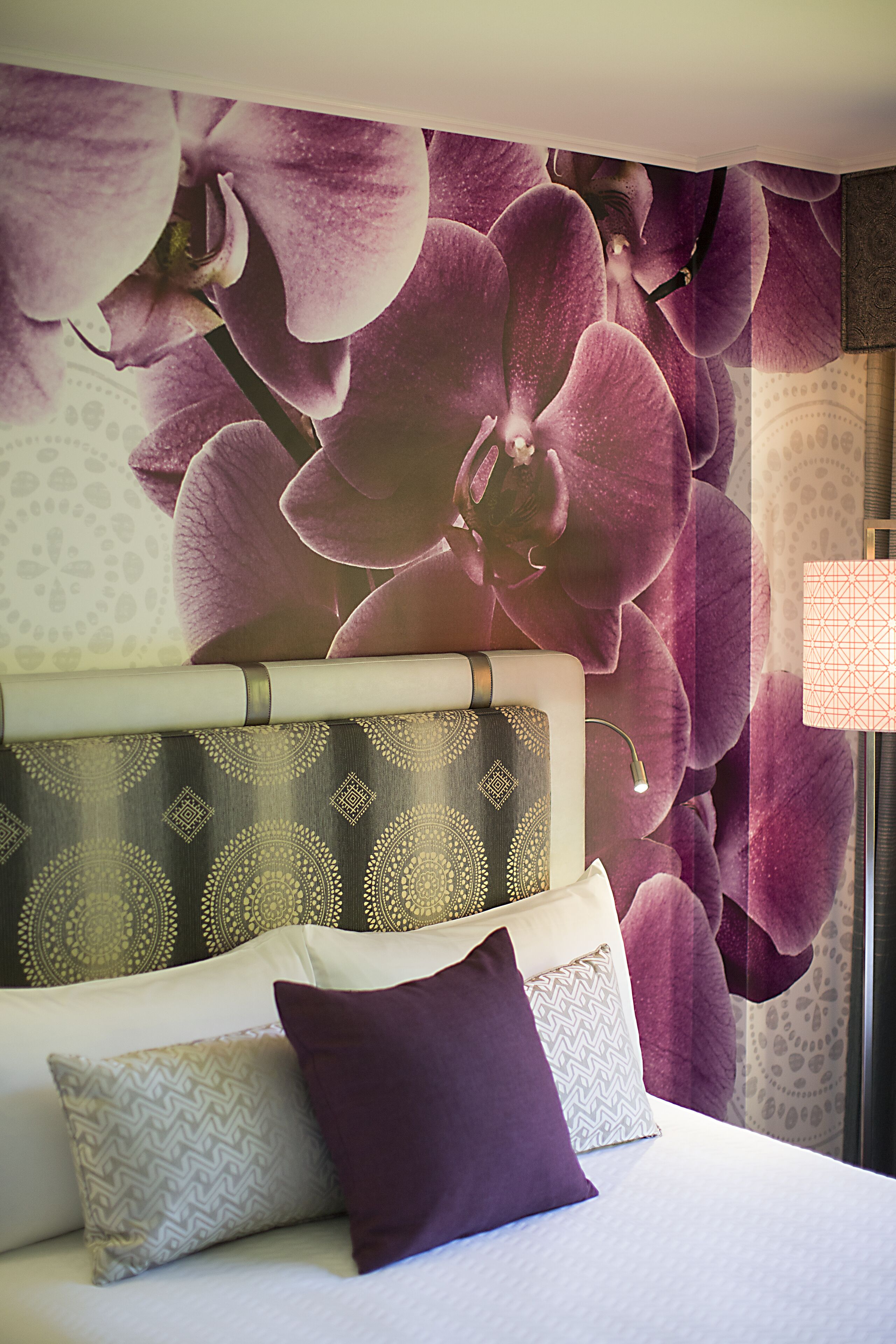 Une chambre à coucher avec un grand décor mural représentant des orchidées, une tête de lit à motifs et de nombreux coussins.
