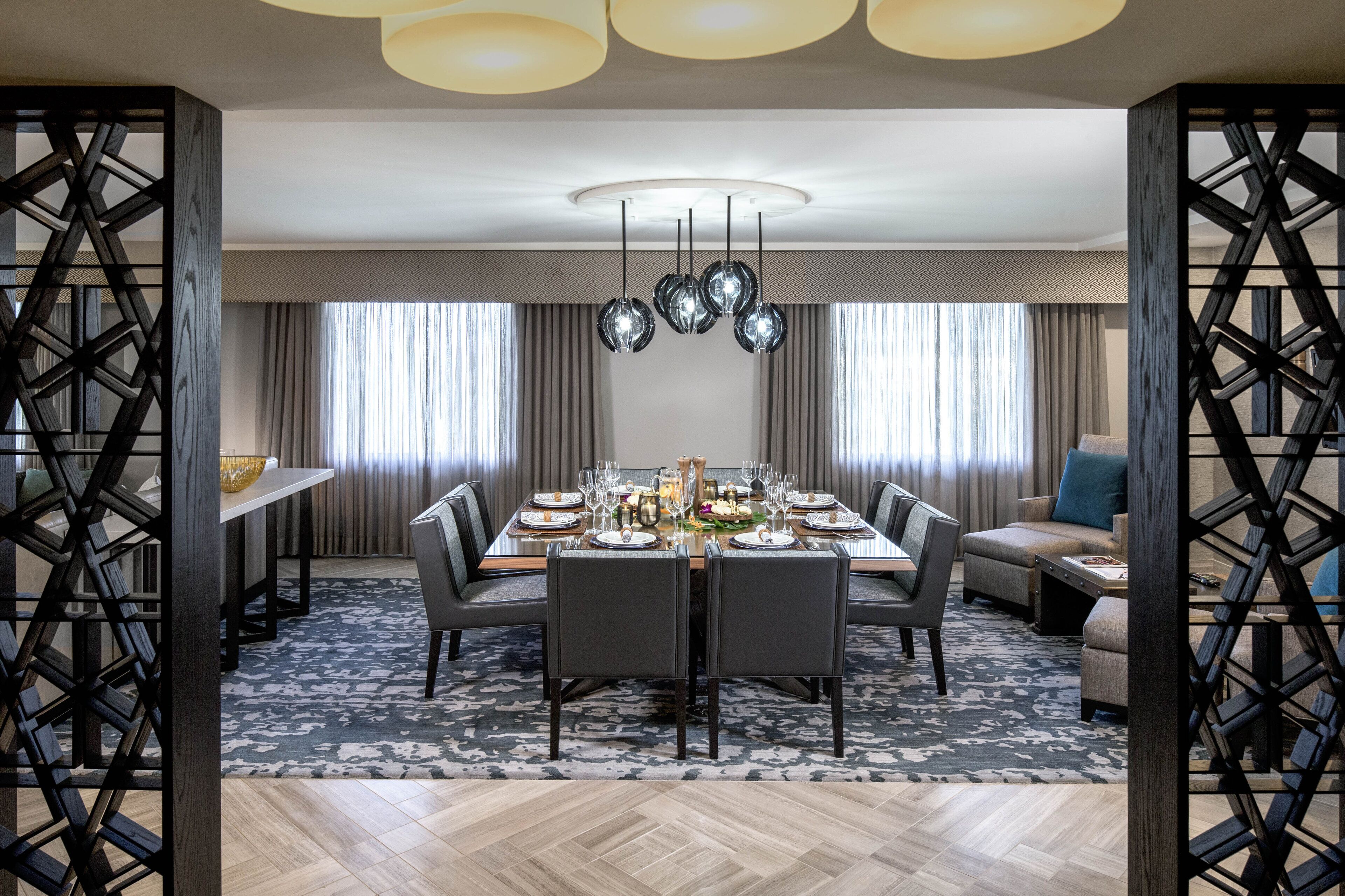 A dining area with a rectangular table set for six, surrounded by chairs, with a patterned rug and a ceiling light fixture above.
