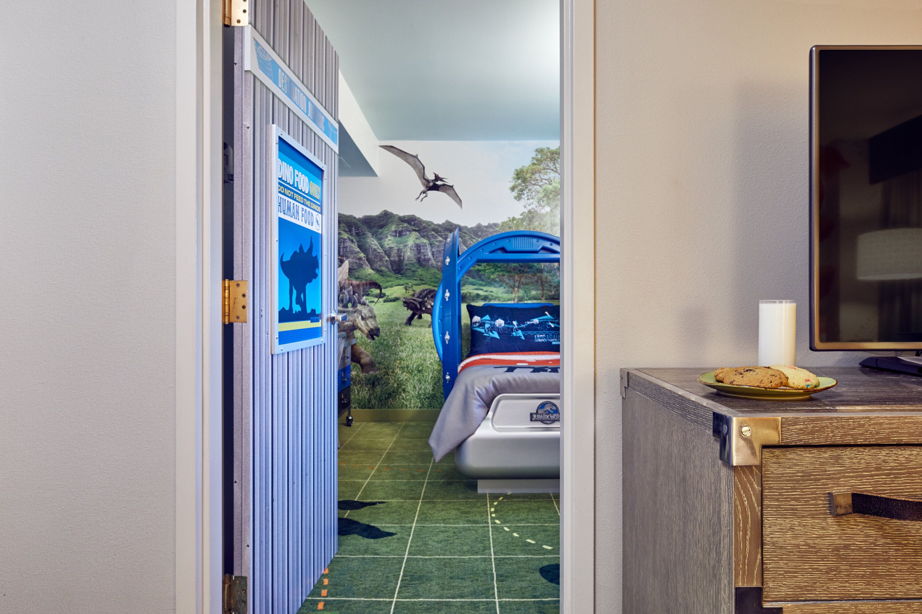 A child's bedroom with a dinosaur-themed mural, a bunk bed, and a small kitchenette area with a plate of cookies.