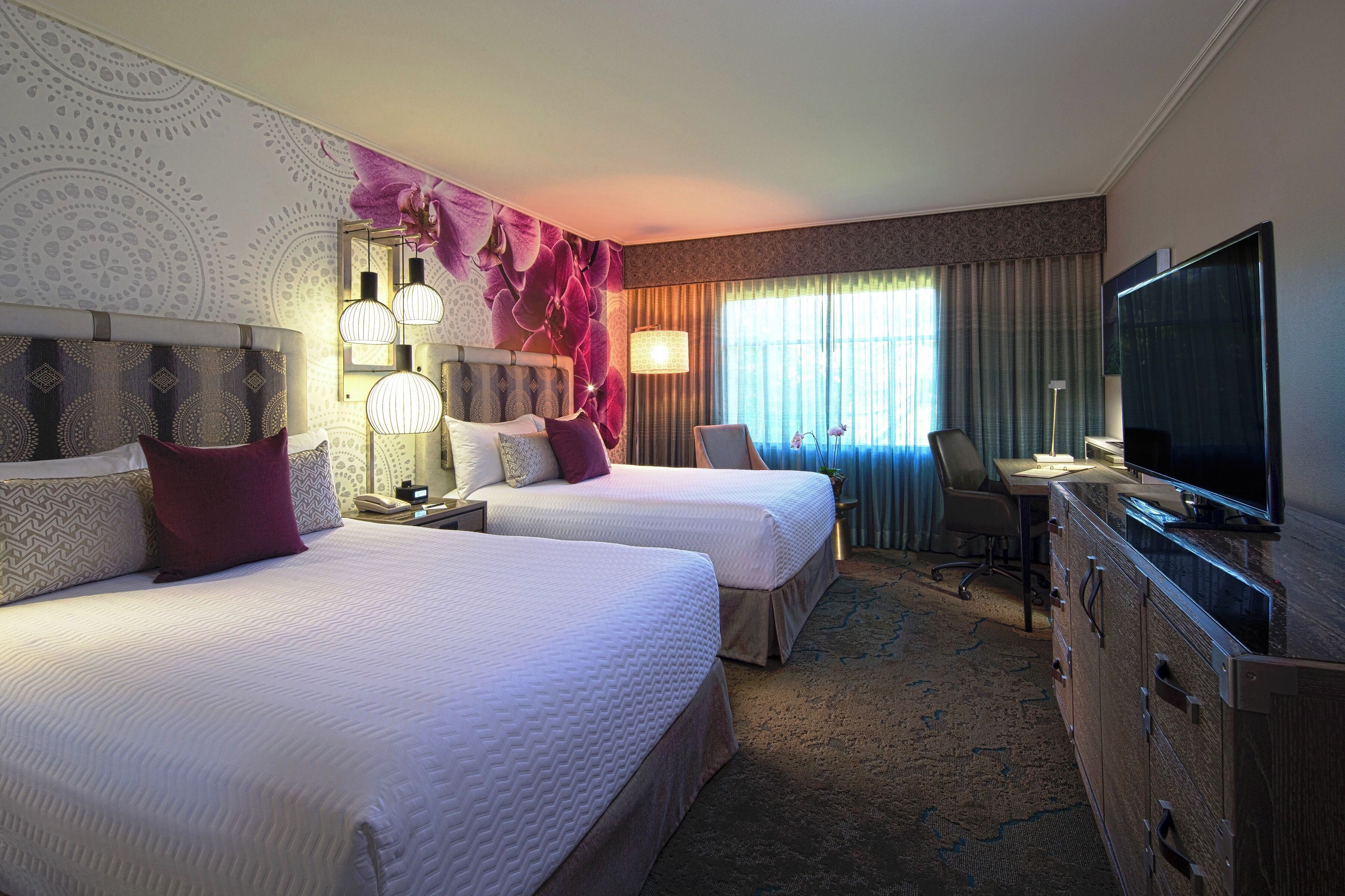 A hotel room with two beds, a desk, a television, and a large window with curtains.