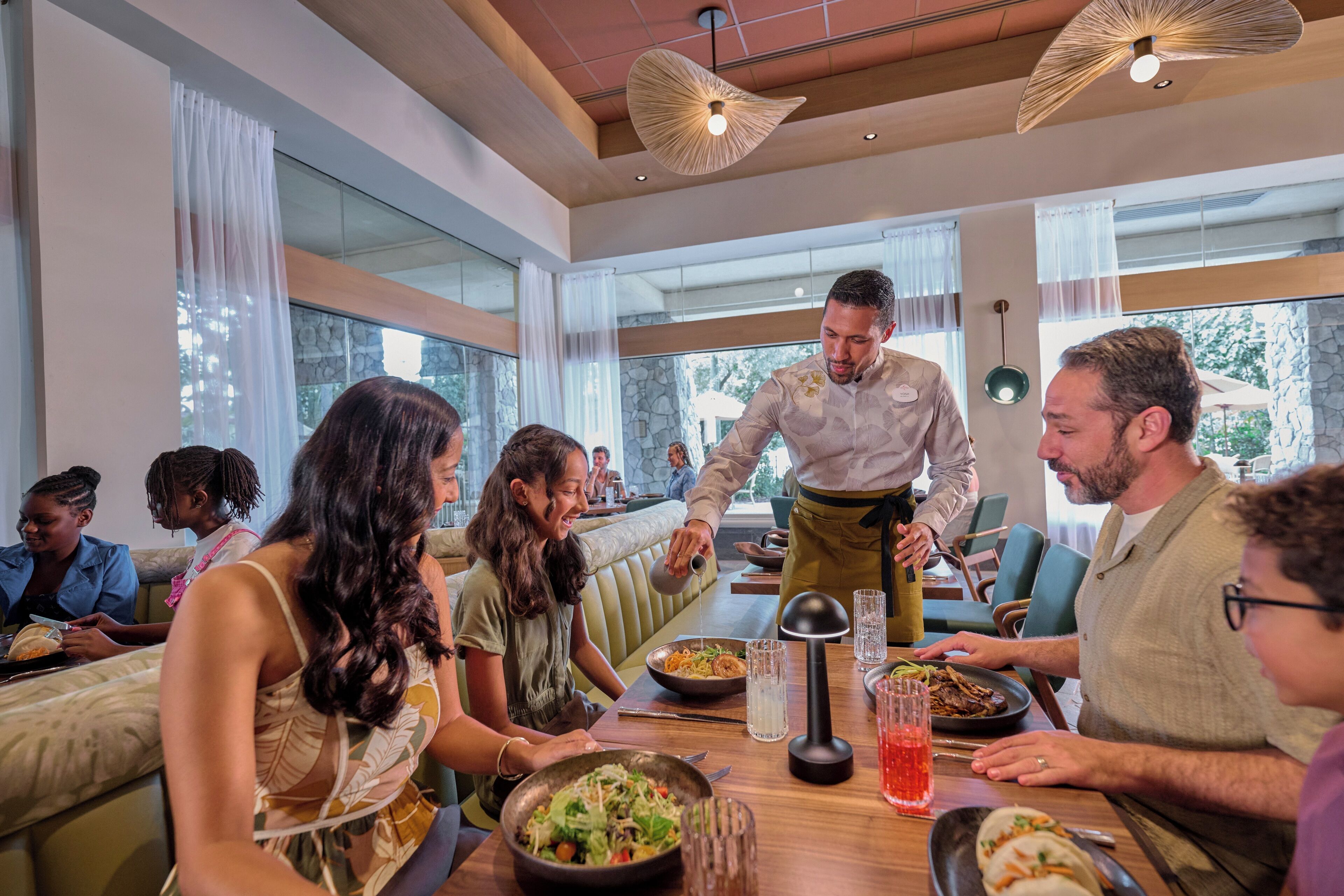 Un restaurant doté d’une longue table commune, où de nombreux clients sont assis, et où un serveur apporte les plats.
