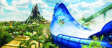 A water park with a large blue water slide, a mountain-like structure, and lush greenery.