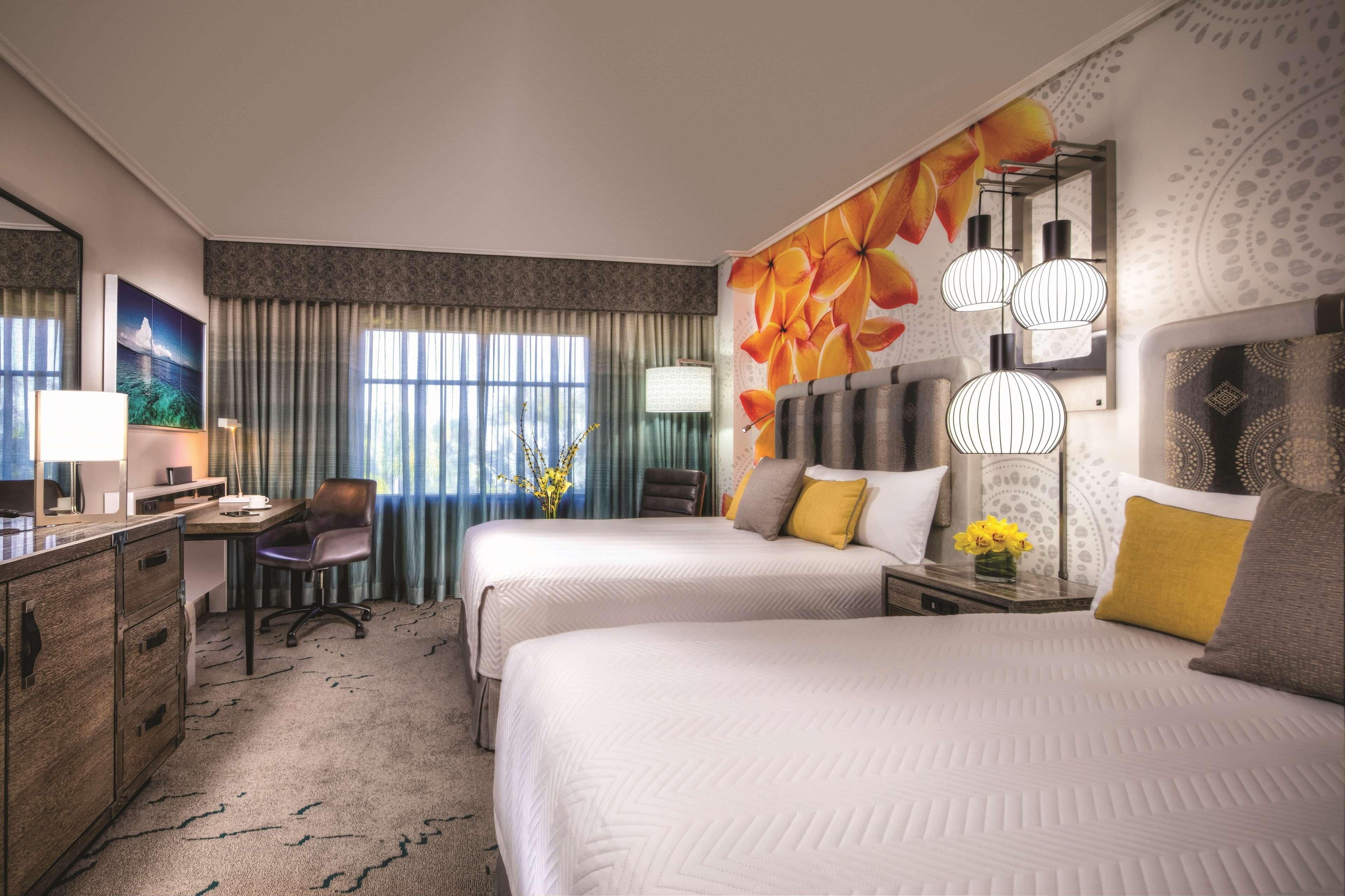 A hotel room with two beds, a desk, a chair, a lamp, and a large window with curtains.