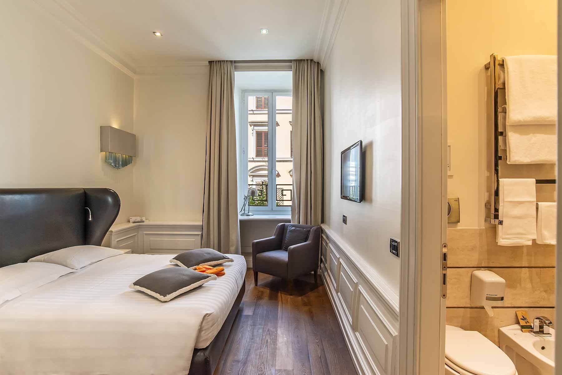 Ett hotellrum med en stor säng, en stol, en TV och ett badrum med handfat och toalett.
