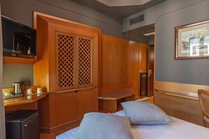 A compact hotel room with a bed, two pillows, a small desk with a kettle, a television, and a wardrobe with a lattice design.