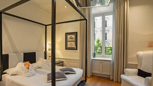 Una cama con dosel, ropa de cama blanca y almohadas grises, un sillón blanco, una mesita de noche con lámpara y una ventana con cortinas.