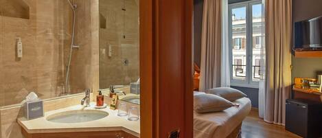 A hotel room with a bathroom featuring a large mirror, a sink, and a shower. The room has a bed, a window with curtains, and a TV on a stand.