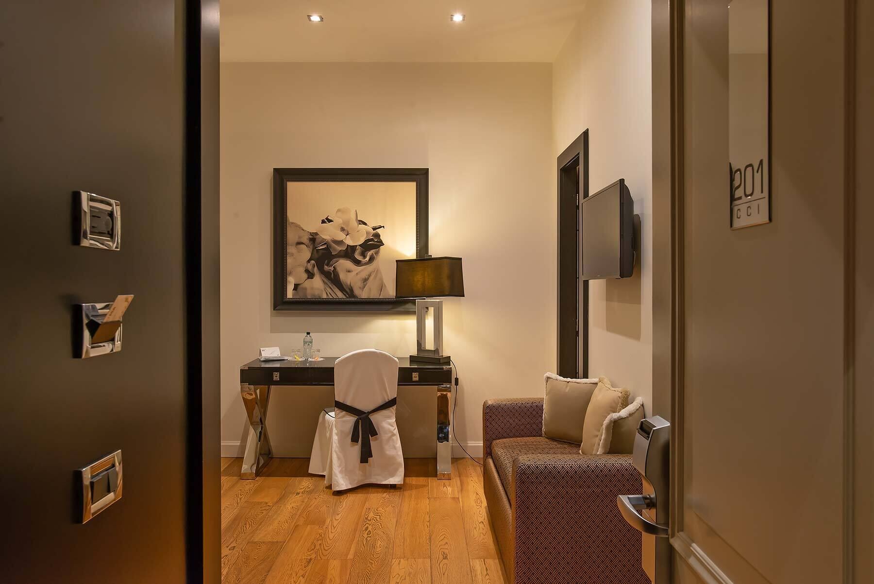 Habitación de hotel con piso de madera, un escritorio con lámpara, una silla, un sofá y un cuadro en la pared.