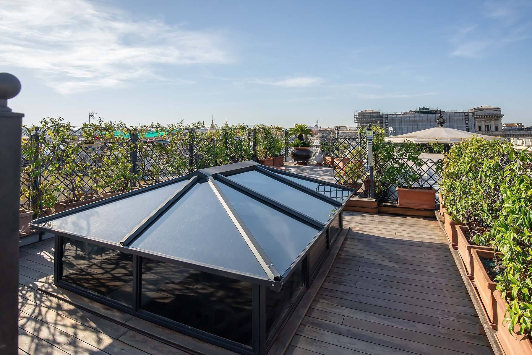 Una terraza en la azotea con techo de vidrio, cubierta de madera, plantas en macetas y vistas de la ciudad.