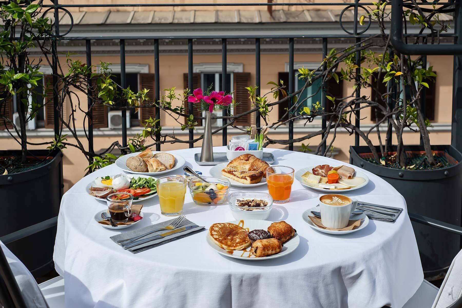 A round table with breakfast items including pancakes, croissants, and a coffee cup, surrounded by potted plants and a balcony railing.
