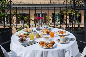 A round table with breakfast items including pancakes, croissants, and a coffee cup, surrounded by potted plants and a balcony railing.