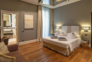A modern hotel room with a large bed, a sitting area with a sofa, a bathroom with a mirror and sink, and a view of the outside through a window.