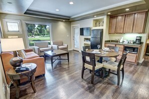 A spacious living area with a sofa, coffee table, and dining table. There is a kitchen area with wooden cabinets and a refrigerator.