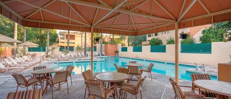 A poolside area with lounge chairs and tables under a canopy, surrounded by a pool and greenery.