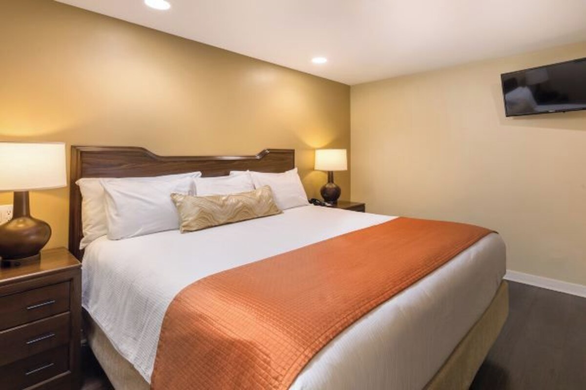Una habitación de hotel con una cama grande, dos mesitas de noche con lámparas y un televisor de pantalla plana montado en la pared.