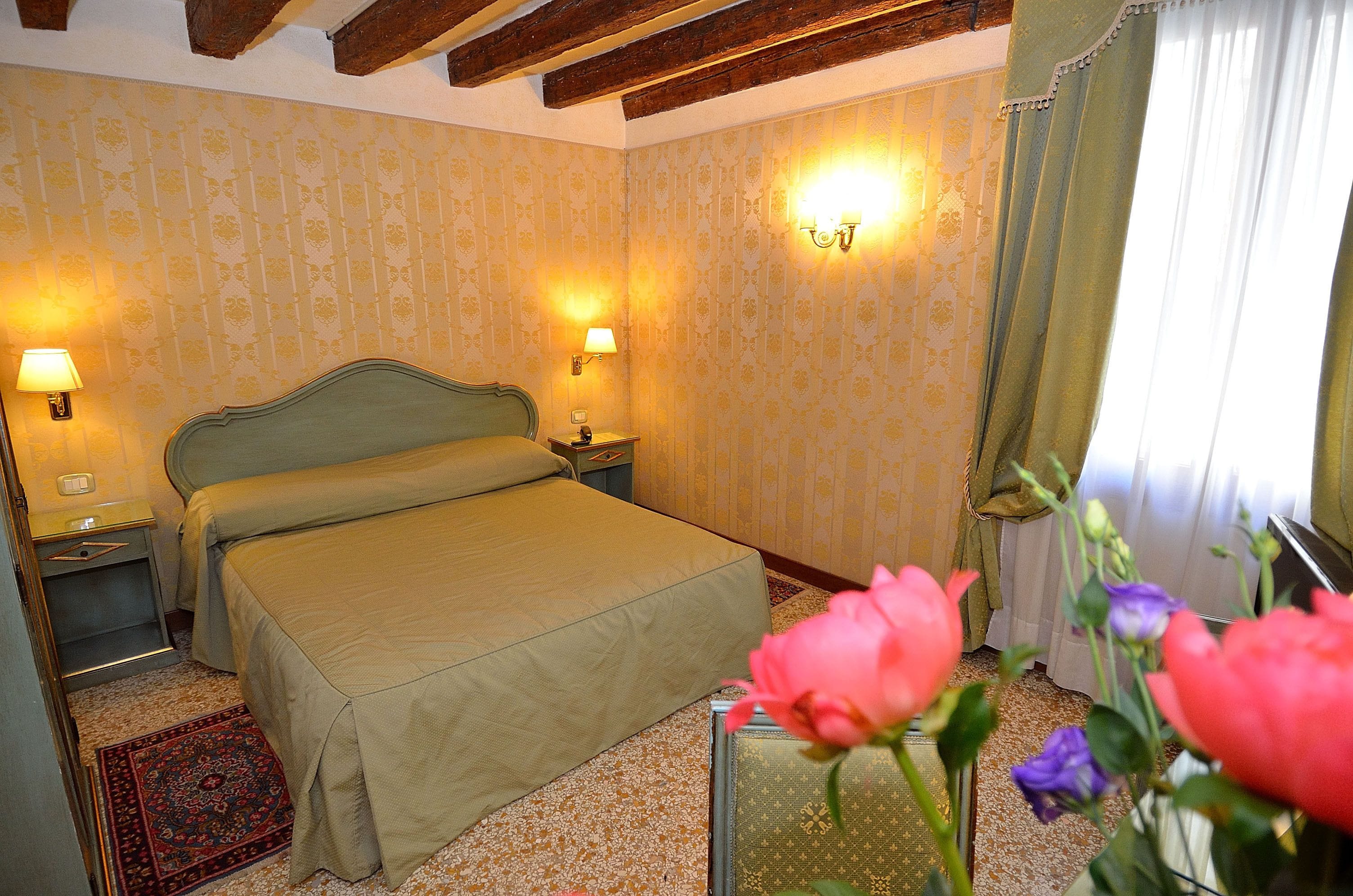 A hotel room with a bed, two bedside lamps, a floral-patterned wallpaper, a window with curtains, and a floral arrangement on the windowsill.