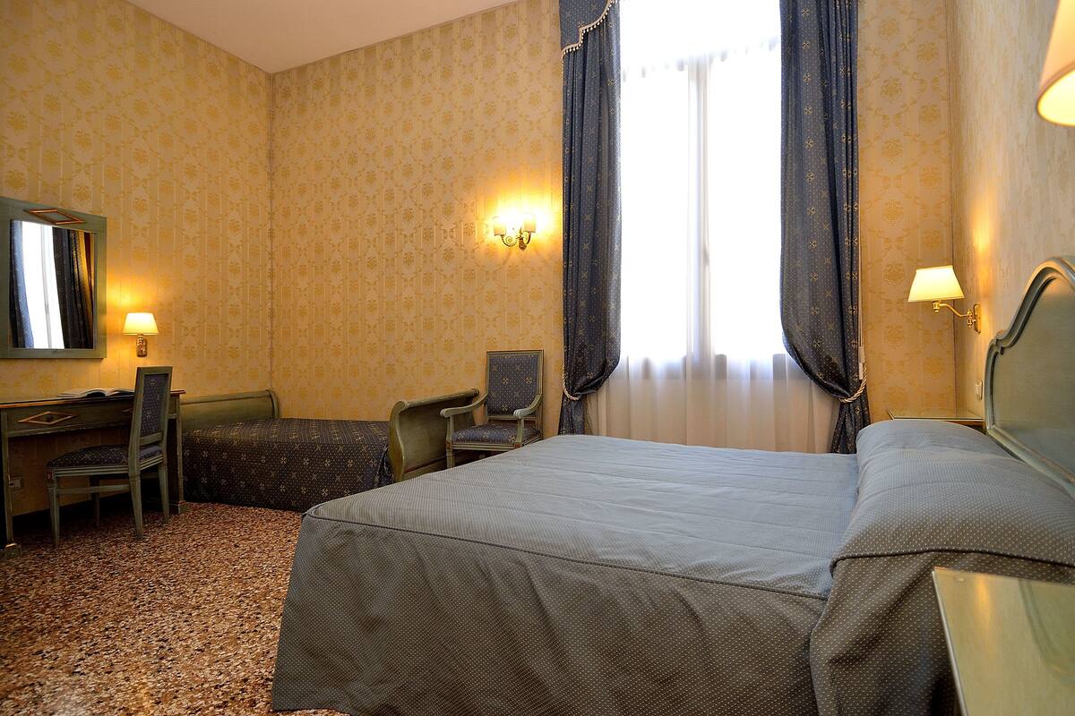 A hotel room with a bed, a desk with a chair, a mirror, and two wall-mounted lamps.