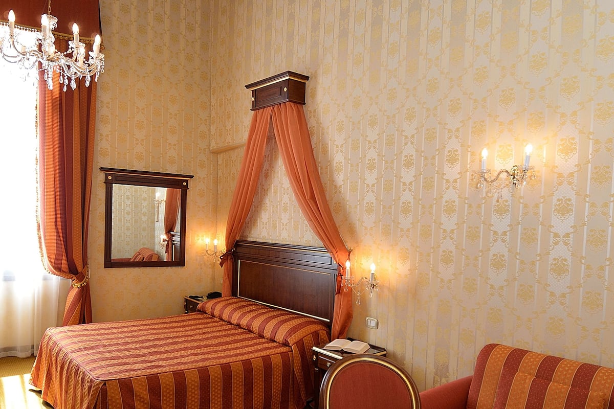 A hotel room with a bed, a sofa, a chair, a table with glasses and a bottle, a mirror, and a chandelier.