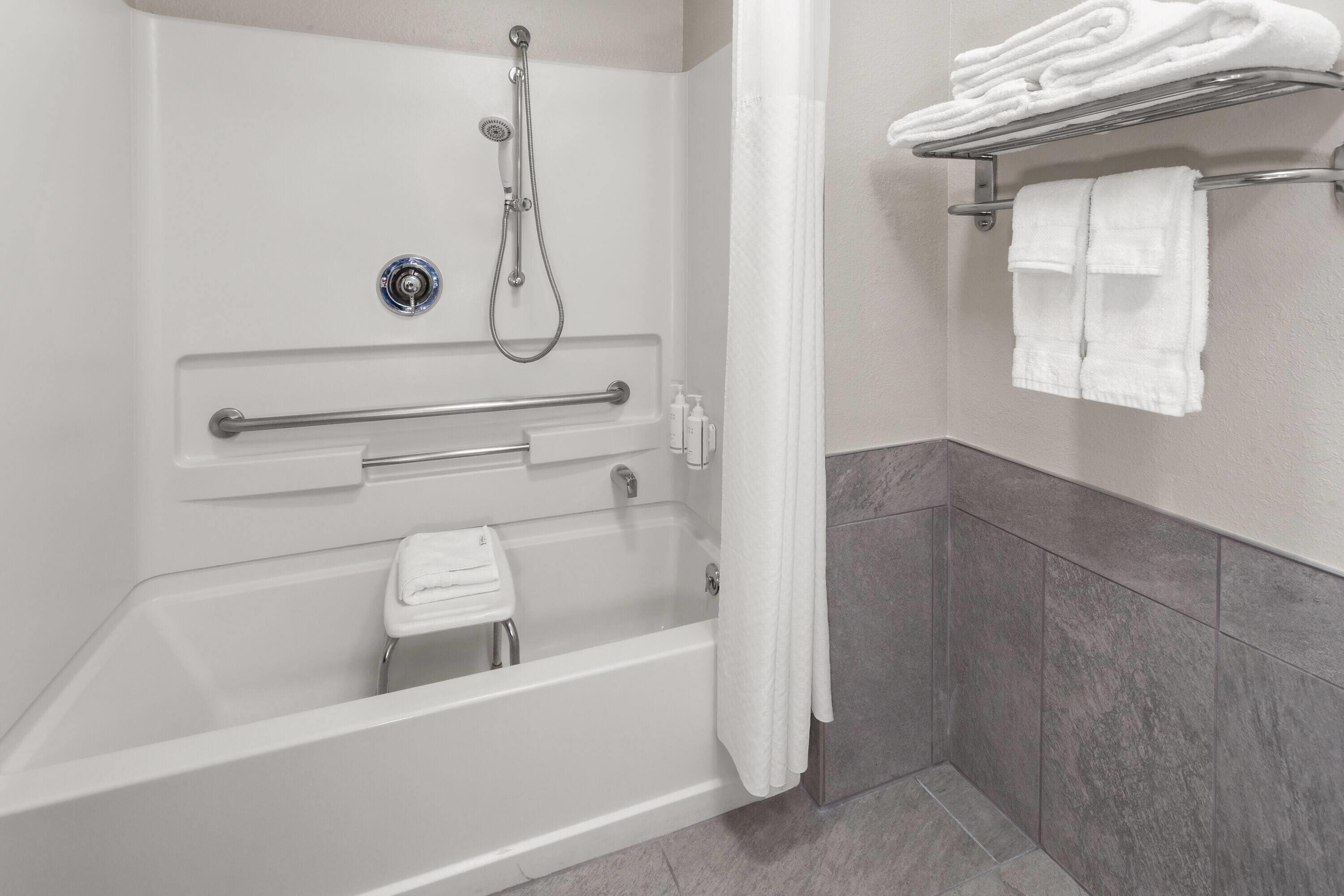 A modern bathroom with a walk-in shower, grab bars, a small stool, and a towel rack.