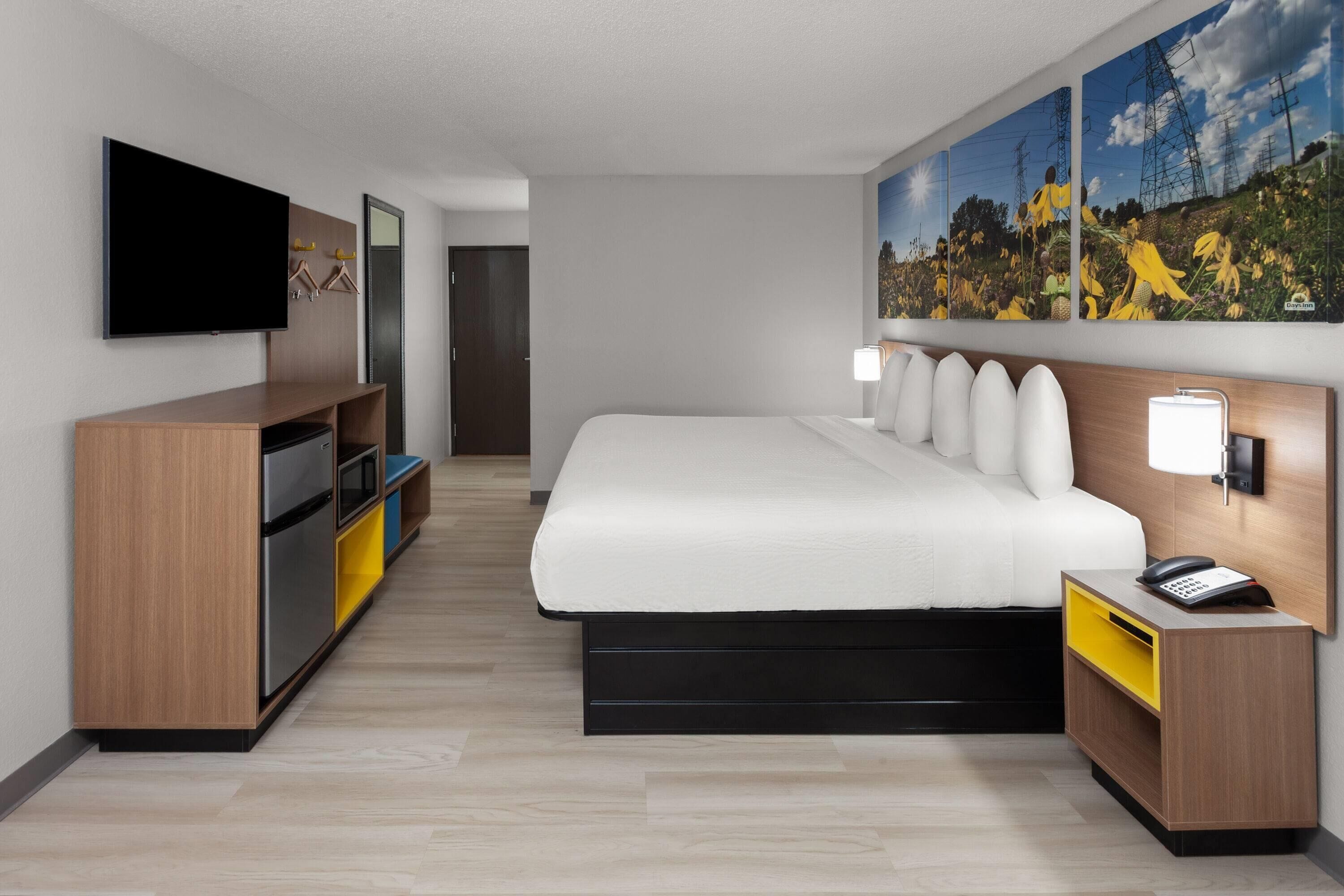 Una habitación de hotel moderna con una cama grande, un televisor de pantalla plana, un tocador de madera y una mesita de noche con lámpara.