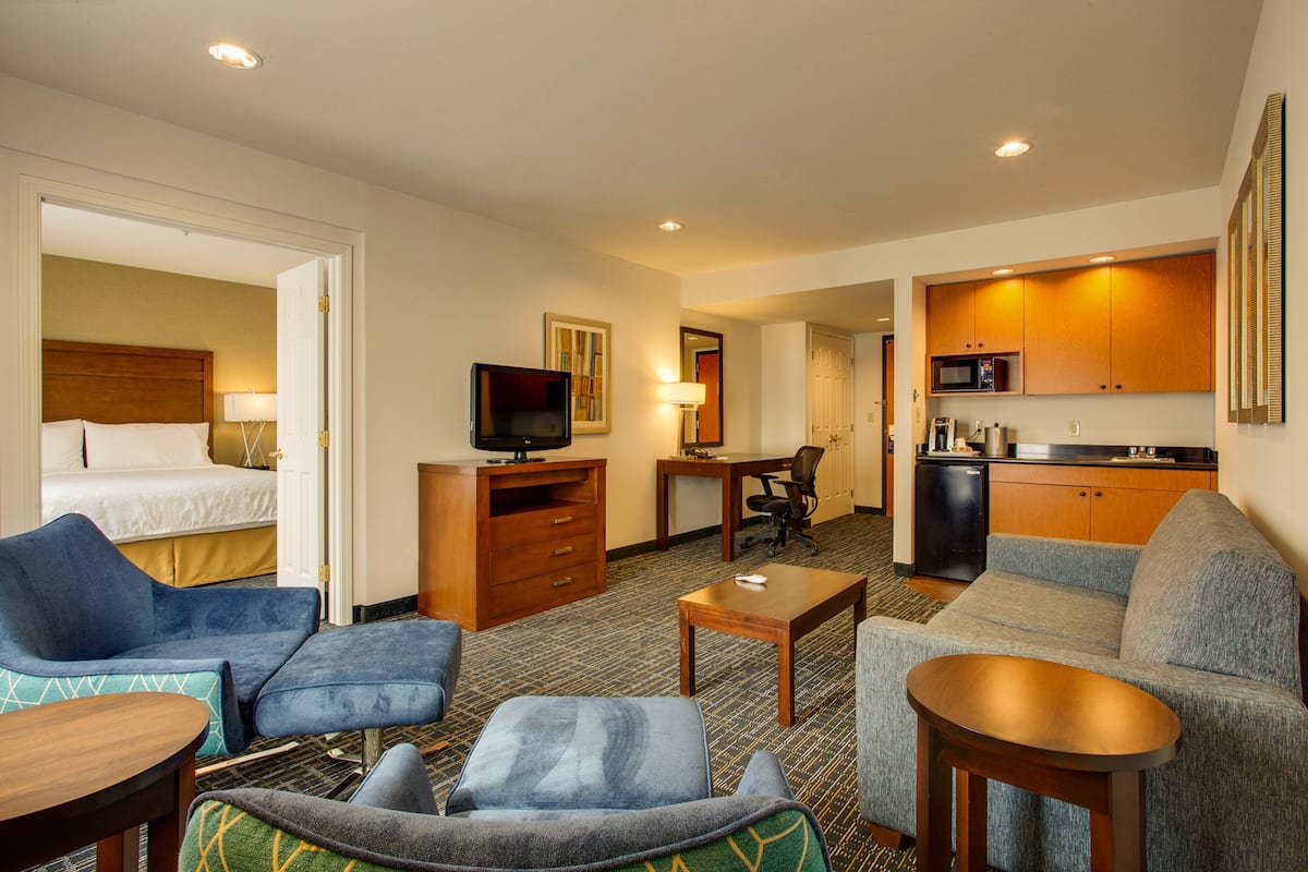 A hotel room with a bed, a TV, a desk, a kitchenette, and a seating area with a sofa and armchairs.