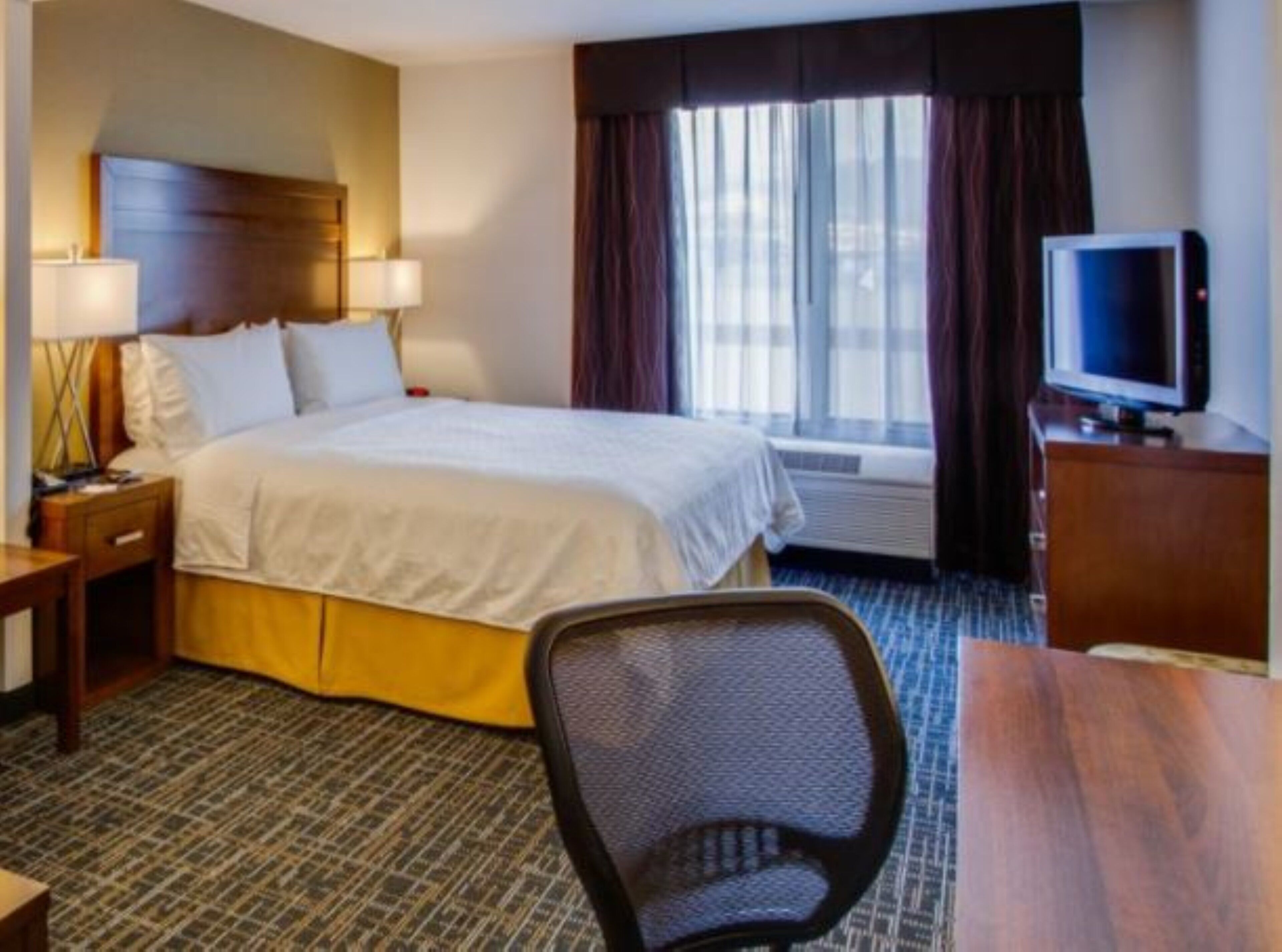 Habitación de hotel con cama, escritorio, silla, televisor y aire acondicionado.