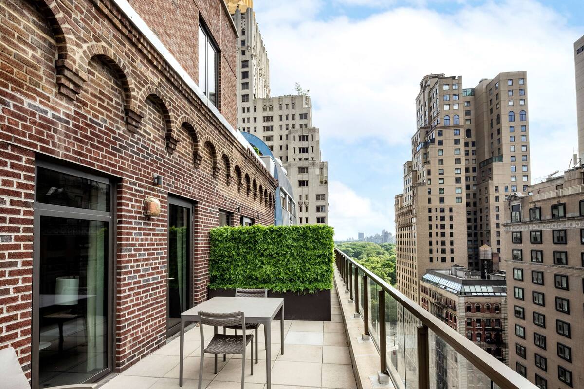 Een balkon met een tafel en stoelen, een groene haag en een stadsgezicht.