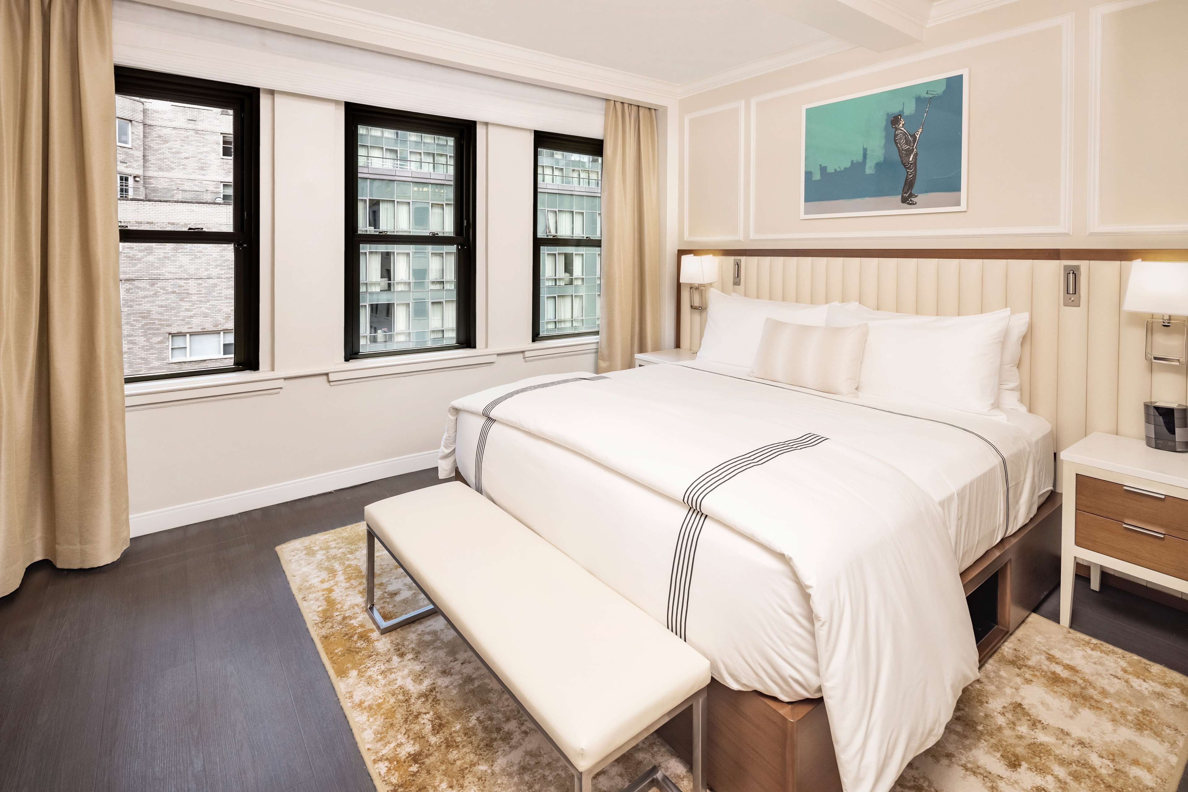 A hotel room with a large bed, a bench, a nightstand, and a painting on the wall.