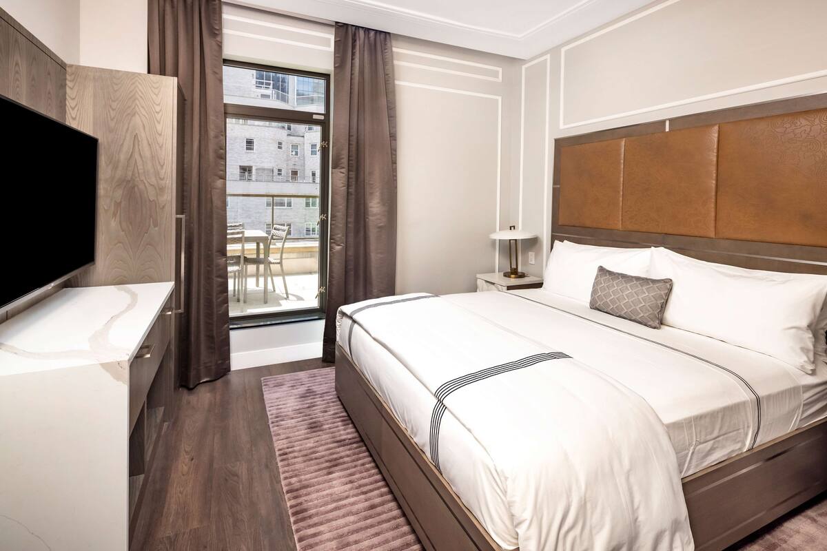Een hotelkamer met een groot bed, een televisie, een raam met gordijnen en uitzicht op gebouwen.