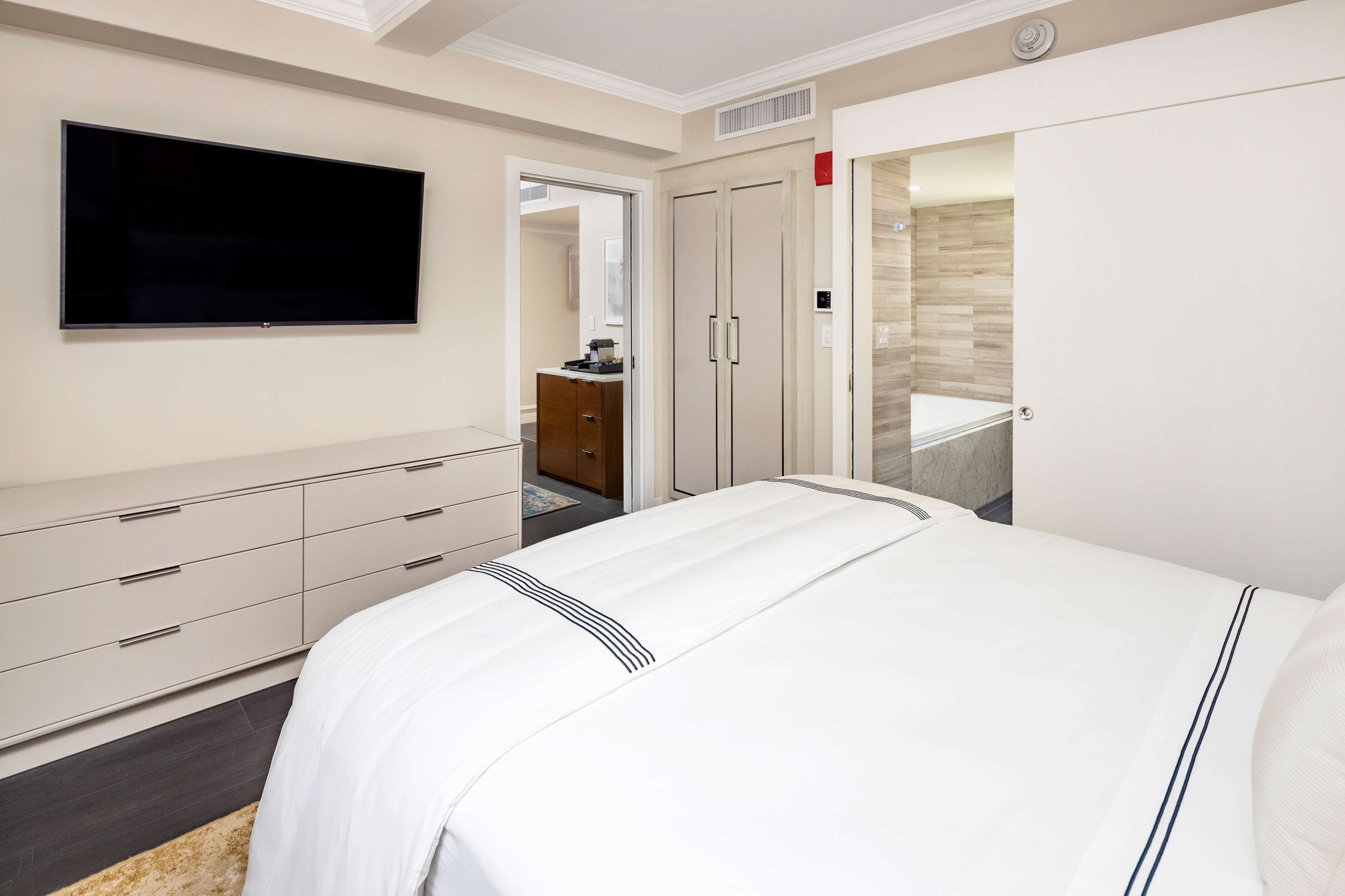 Ein Hotelzimmer mit Bett, Wandfernseher, Kleiderschrank und einem durchgegangenen Tür sichtbaren Badezimmer.
