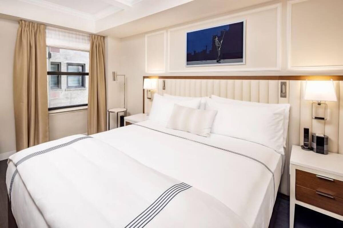 Hotelkamer met een groot bed, nachtkastje, lamp, schilderij aan de muur en raam met gordijnen.