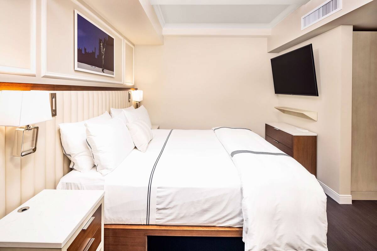 Een hotelkamer met een bed, nachtkastje, lamp, televisie en kunst aan de muur.