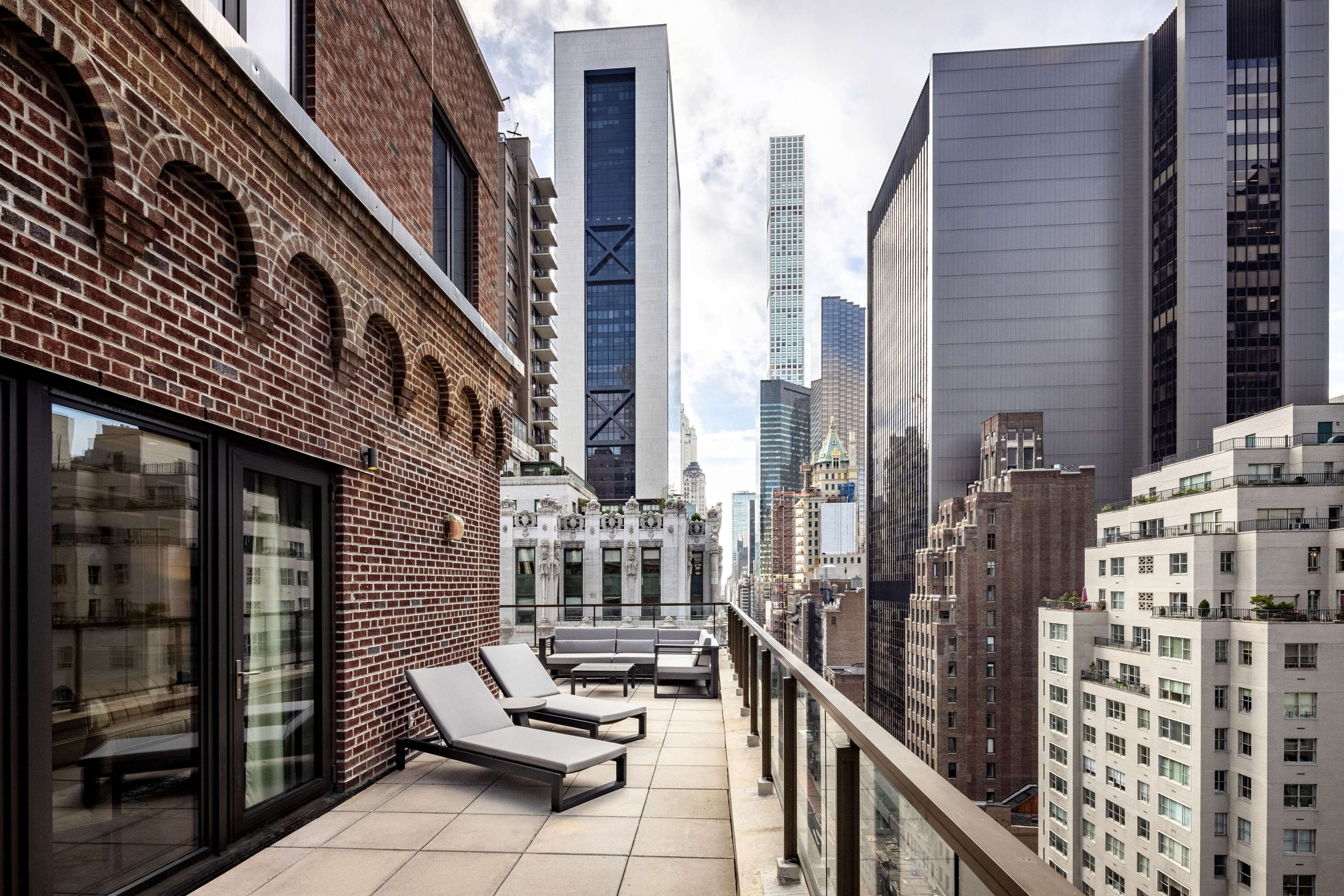 Una terraza en la azotea con sillones de descanso, un edificio de ladrillo y modernos rascacielos al fondo.