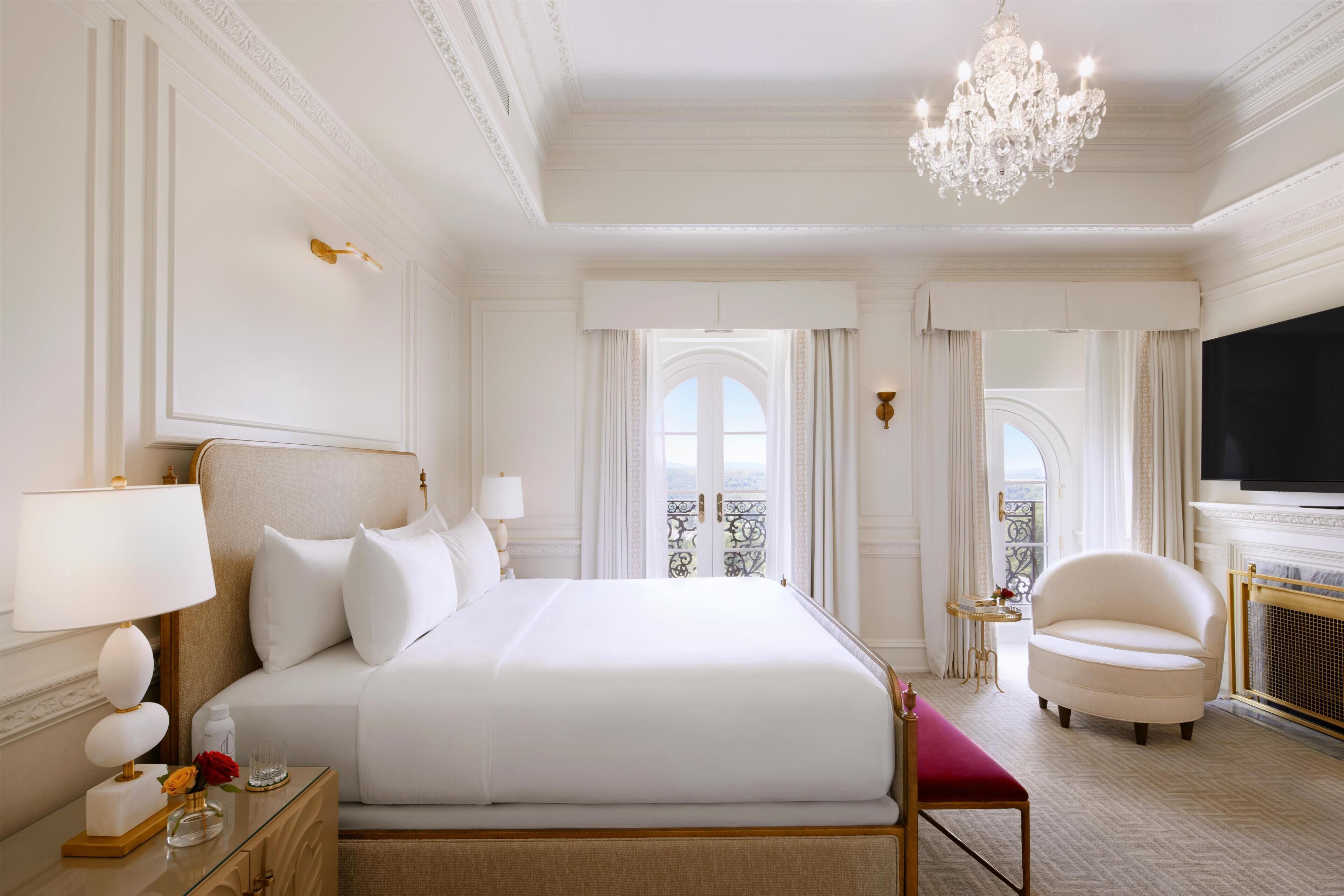 A hotel room with a large bed, a chandelier, a television, and a view of the outdoors.