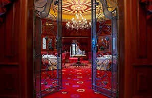 A grand hallway with ornate metal gates, a chandelier, and a red carpet leading to a dining area with tables and chairs.