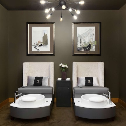 A spa room with two massage chairs, a central table with a vase of flowers, framed artwork on the walls, and a floor lamp.