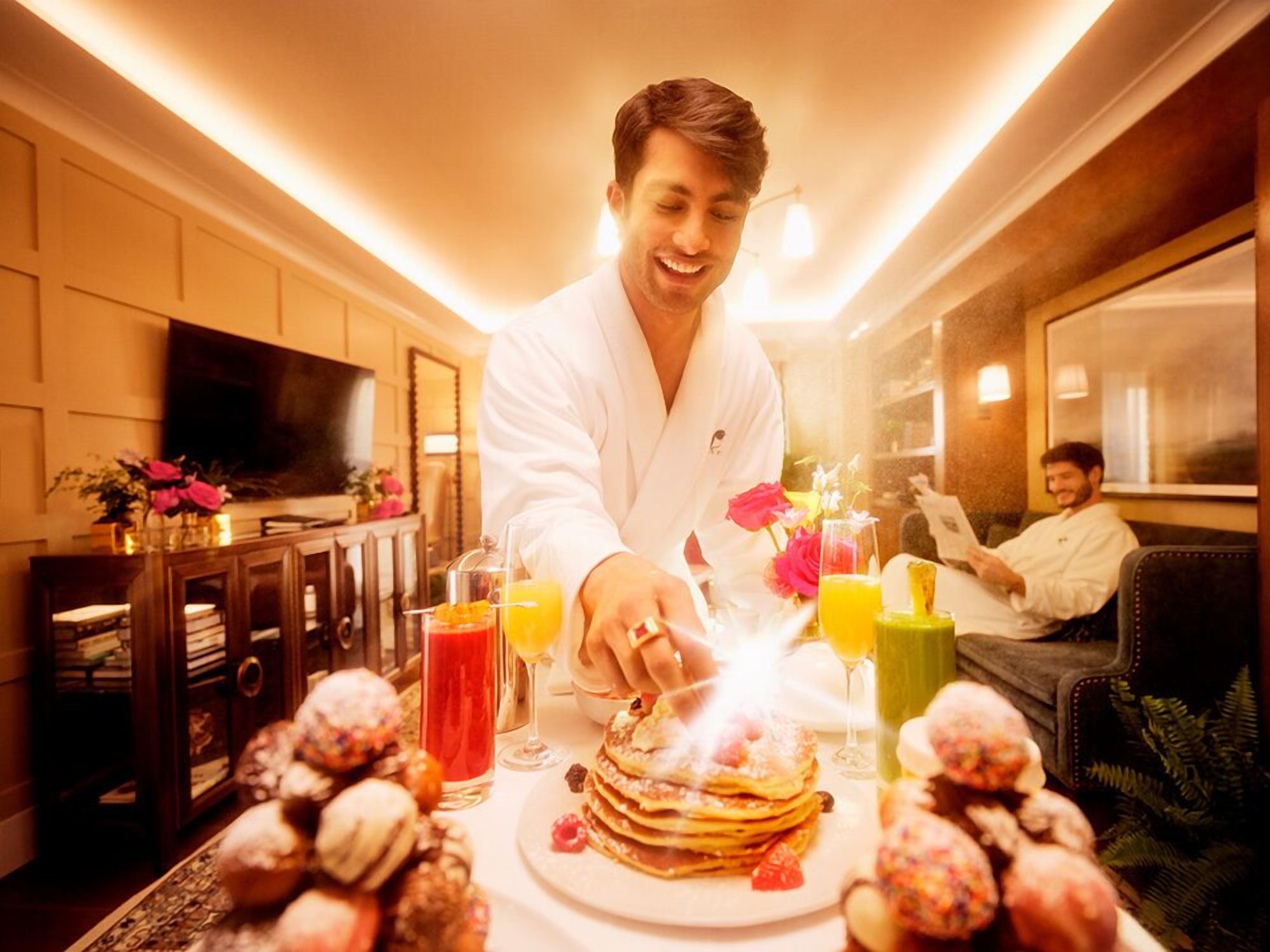 A person serving pancakes with a lit candle on a table in a warmly lit room with a television and a bookshelf.