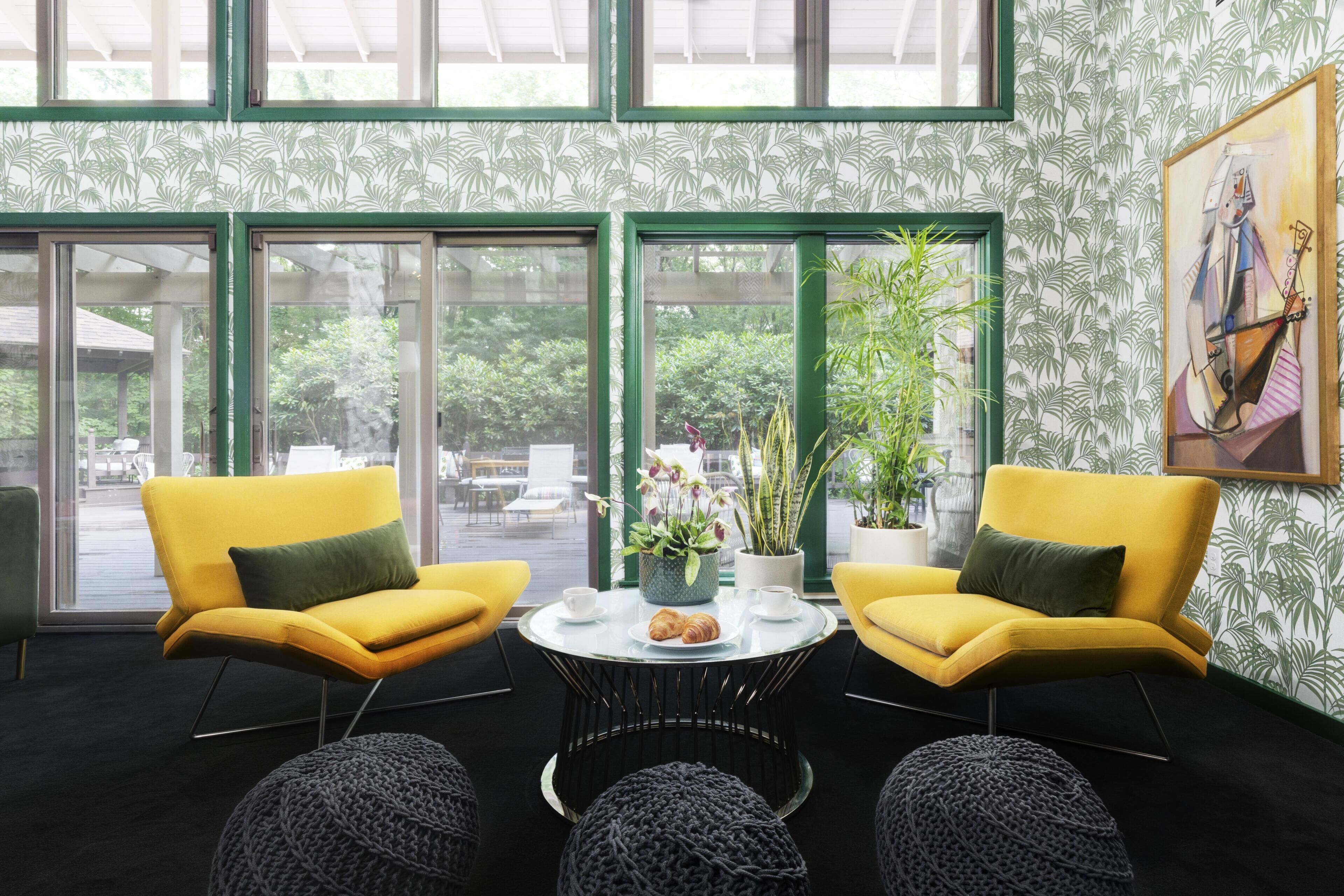 Un intérieur moderne avec deux chaises jaunes, une table ronde agrémentée d’un vase et de fleurs, et de grandes fenêtres offrant une vue sur l’extérieur.