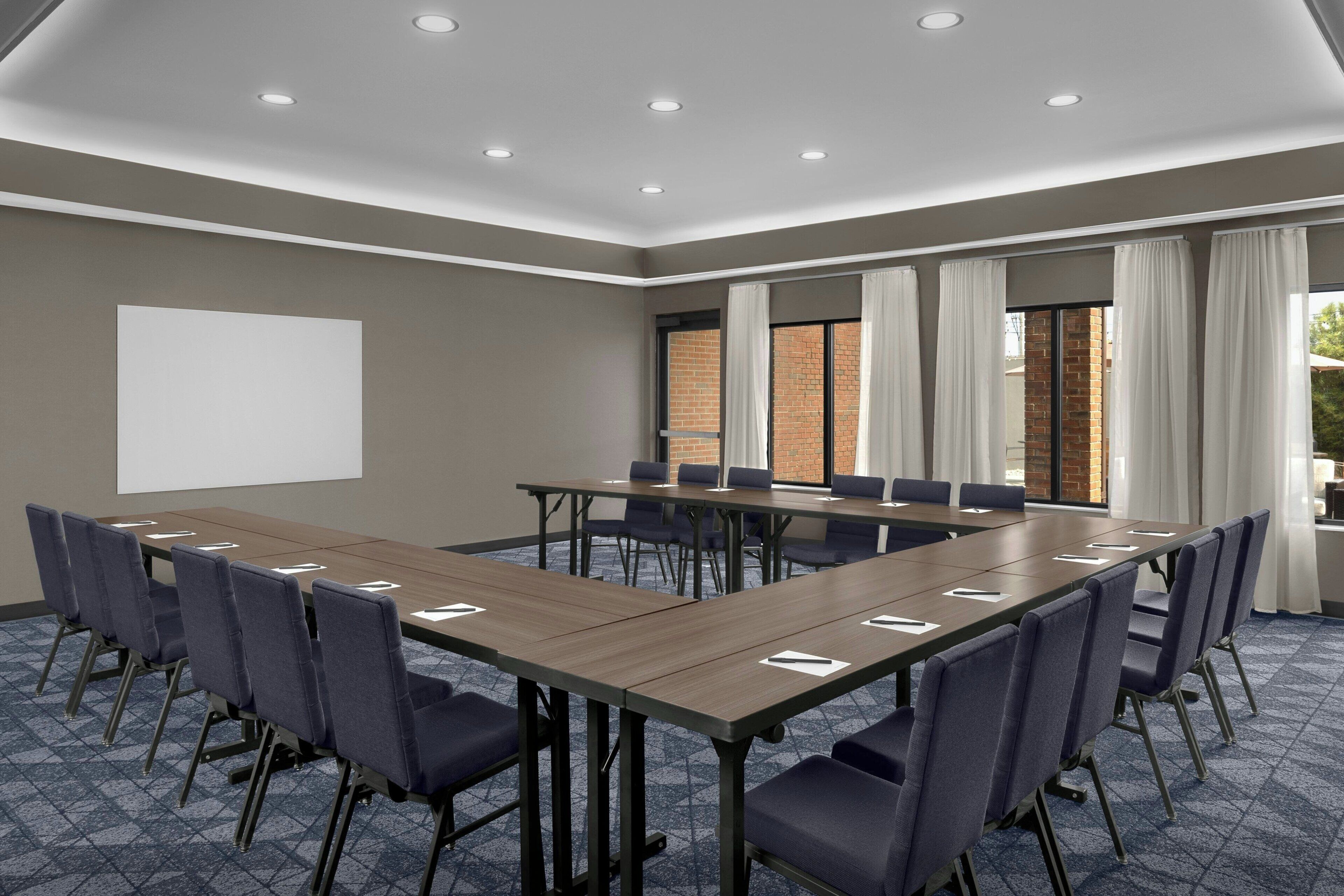 Ein Konferenzraum mit einem langen Tisch, Stühlen, einer Projektionsleinwand und Fenstern mit Vorhängen.