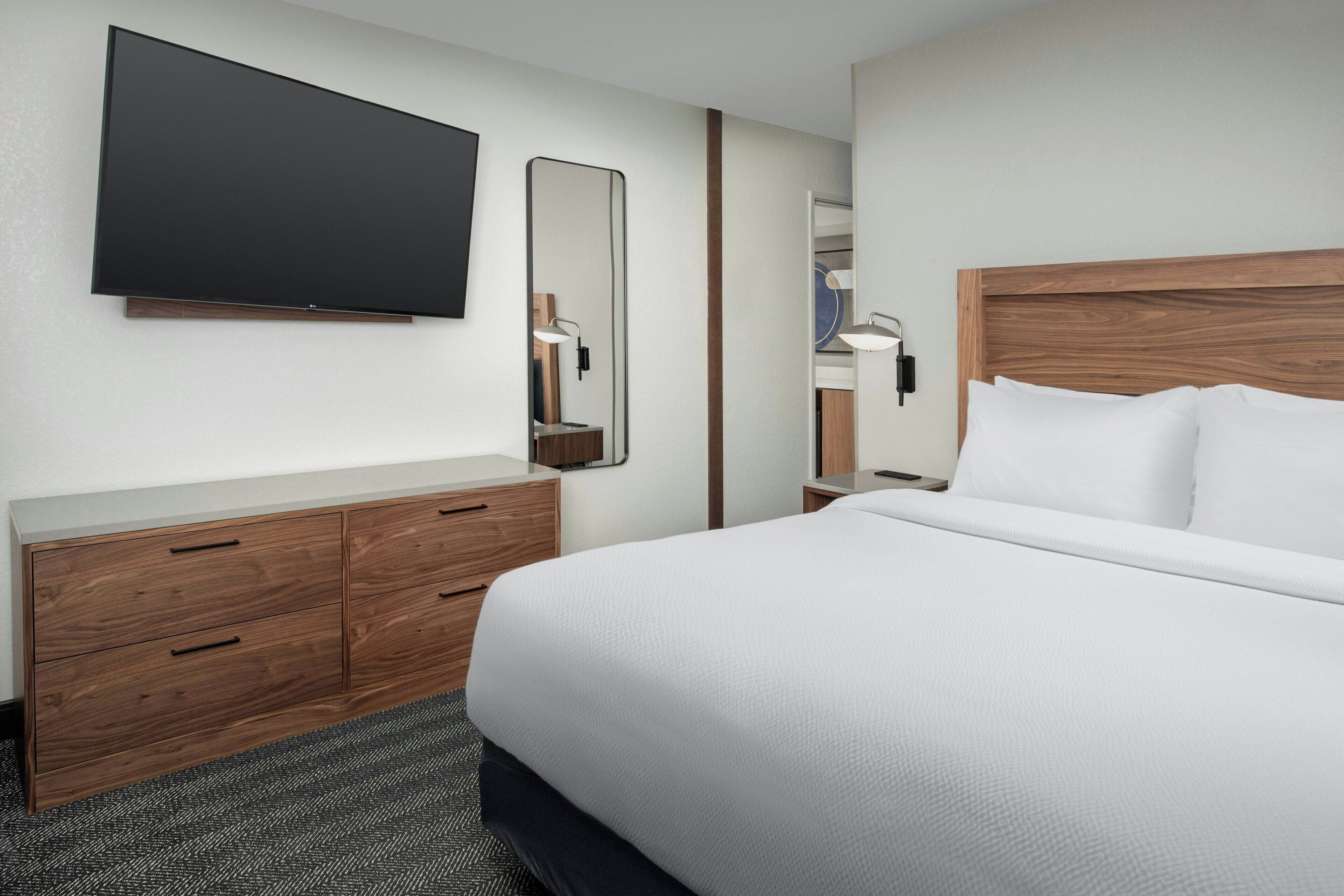 Una habitación de hotel con una cama grande, un aparador de madera, un televisor de pantalla plana, un espejo y una mesita de noche con lámpara.