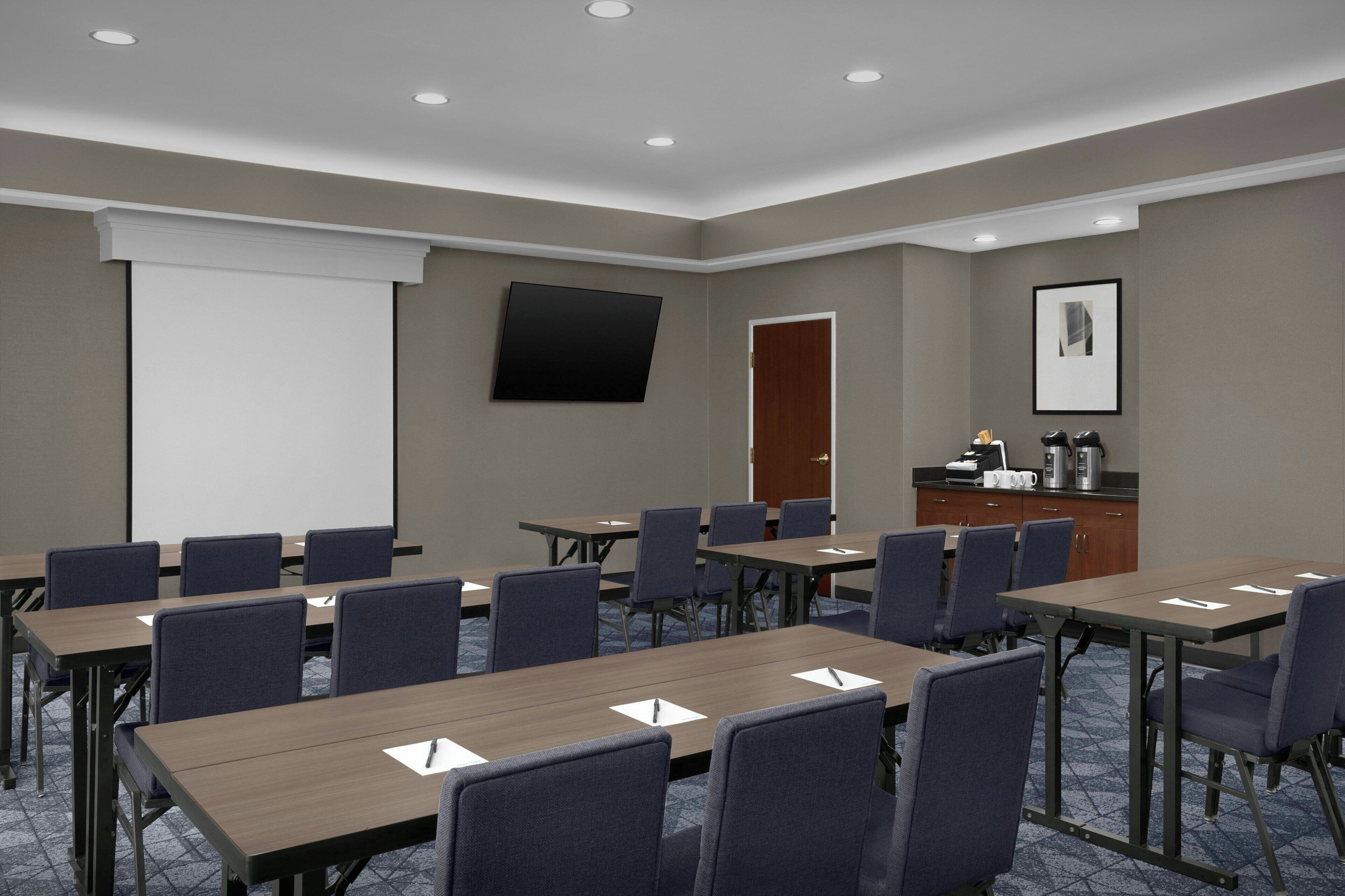 A conference room with multiple tables and chairs, a projector screen, a door, and a coffee station.