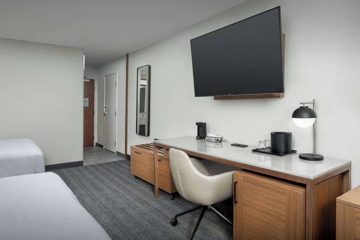 Habitación de hotel con cama, escritorio con silla, televisor de pantalla plana, espejo y lámpara.