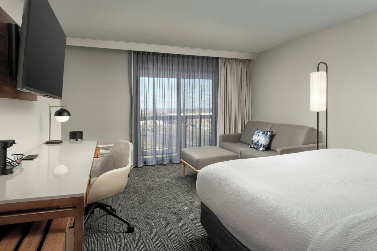 Habitación de hotel con cama, escritorio, silla, sofá, televisor, lámpara y ventana con cortinas.