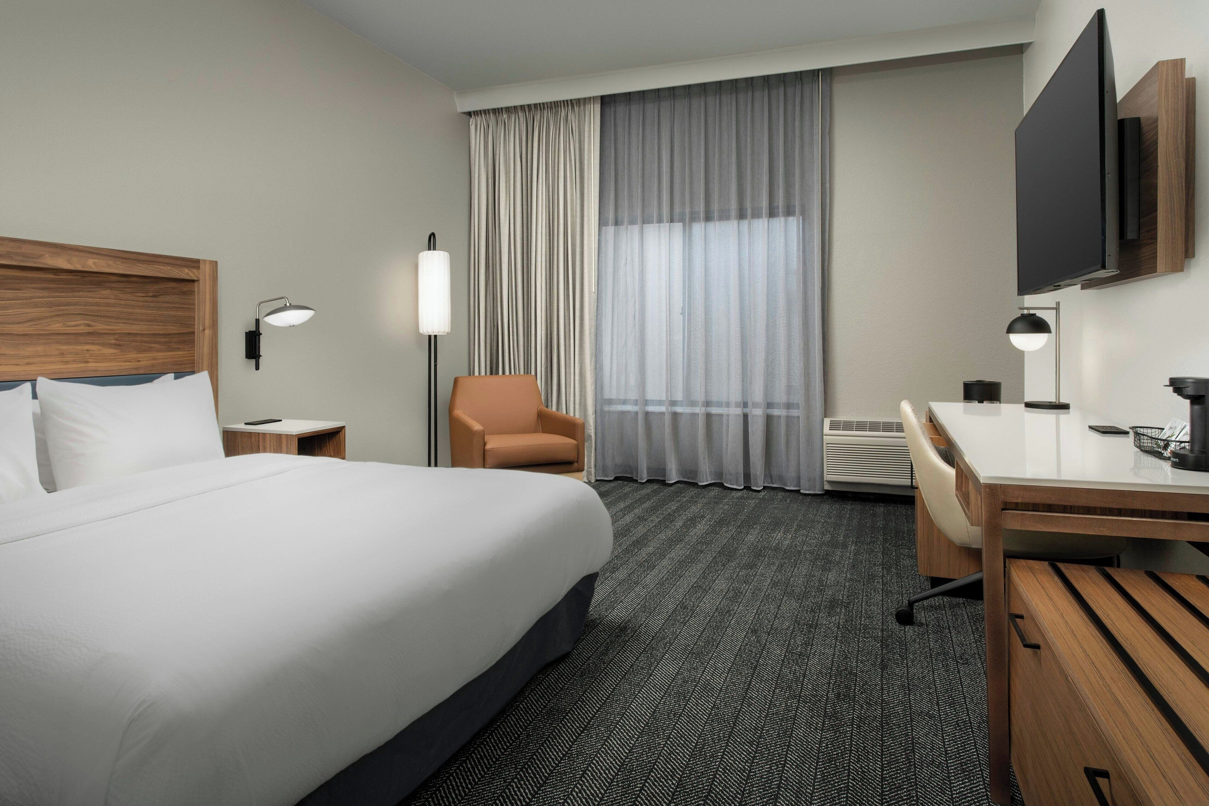 Habitación de hotel con una cama grande, un escritorio, una silla, un televisor y una ventana con cortinas.